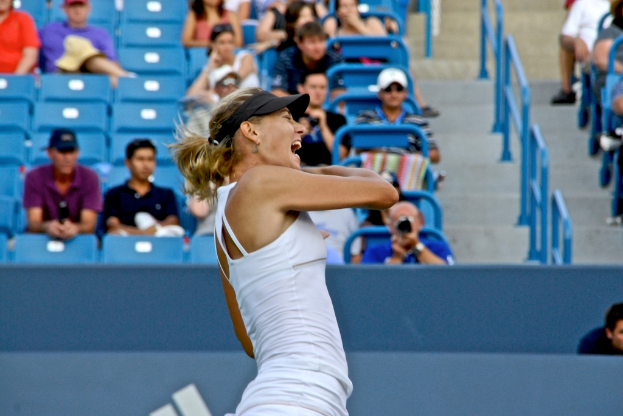 Eine Frau in einem weißen Oberteil und schwarzer Mütze steht vor einem blauen Stadionplatz mit sitzenden Zuschauern.
