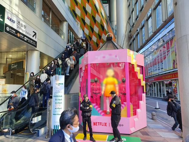 Eine Gruppe von Menschen steht vor einem Gebäude mit Glaswänden, Säulen und Texttafeln, einige tragen Masken, während eine Rolltreppe mit Fahrgästen zu sehen ist; ein pinker Kasten in der Mitte zeigt Schaufensterpuppen und trägt die Aufschrift "Netflix's neuer Netflix-Stand in Tokio."