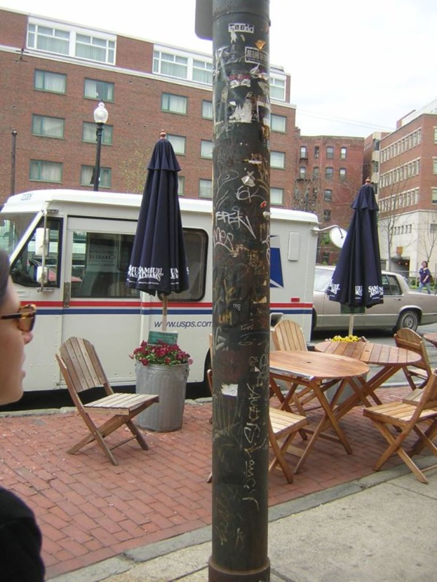 Ein Tisch und Stühle sind auf einem Gehweg aufgestellt, mit Autos und Lieferwagen, die auf der benachbarten Straße geparkt sind, und Gebäuden im Hintergrund.