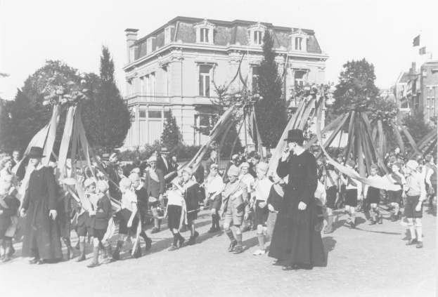 Schwarzes und weißes Bild einer Gruppe von Menschen, die eine Straße entlanggehen und Fähnen halten, mit Hüten, Bäumen, Gebäuden und einem Maibaum im Hintergrund.