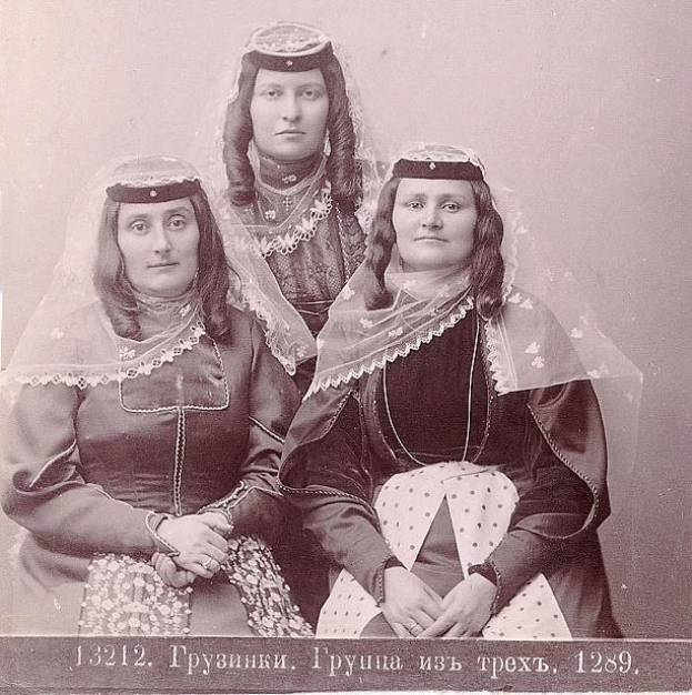 Drei Frauen in traditionellen russischen Hochzeitskleidern mit farbenfroher Stickerei und Perlen stehen vor einer weißen Wand in einem Schwarz-Weiß-Foto, das als russisches Hochzeitsfoto bezeichnet wird.