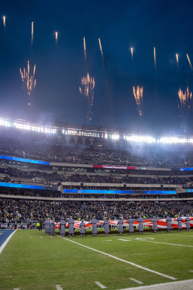 Ein Fußballstadion voller stehender Zuschauer unter hellen Lichtern, umgeben von textbedeckten Tafeln, mit Feuerwerk, das am Himmel explodiert.