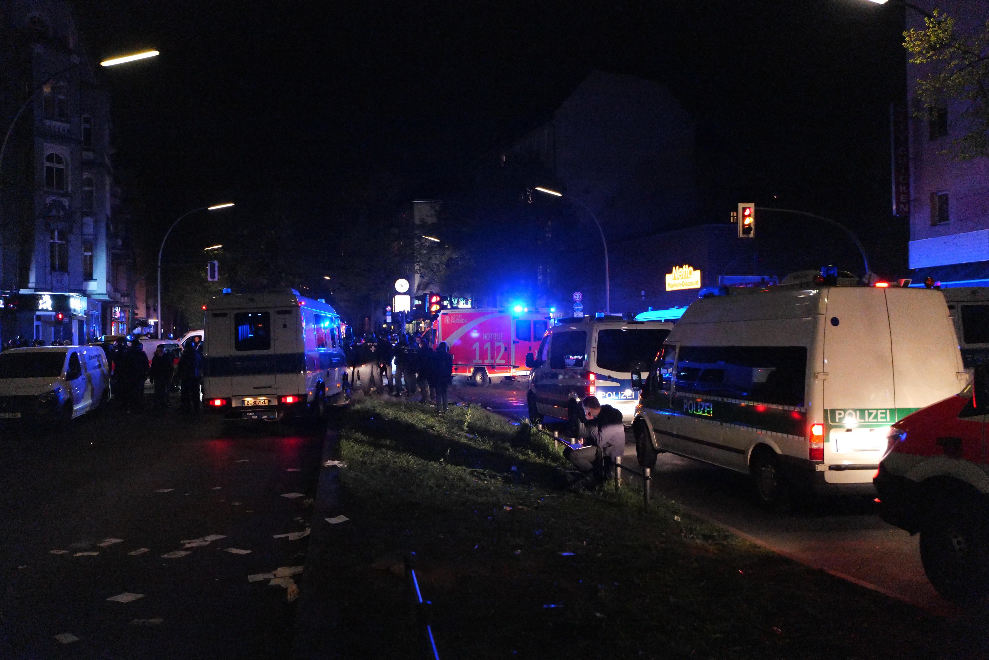 Eine Gruppe von Polizeiwagen steht nachts am Straßenrand, mit Fahrzeugen auf der Straße, Menschen stehen, Gras, Straßenlaternen, Verkehrszeichen, Bäume, Gebäude mit Fenstern und ein dunkler Himmel im Hintergrund.