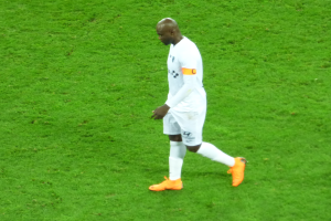 Ein Fußballspieler in einem weißen T-Shirt, weißen Shorts und orangefarbenen Schuhen läuft über ein saftiges grünes Feld.
