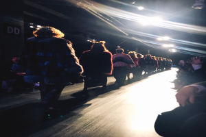 Eine Gruppe von Menschen läuft über einen hell erleuchteten Laufsteg bei einer Fashion Show, mit Zuschauern auf beiden Seiten.