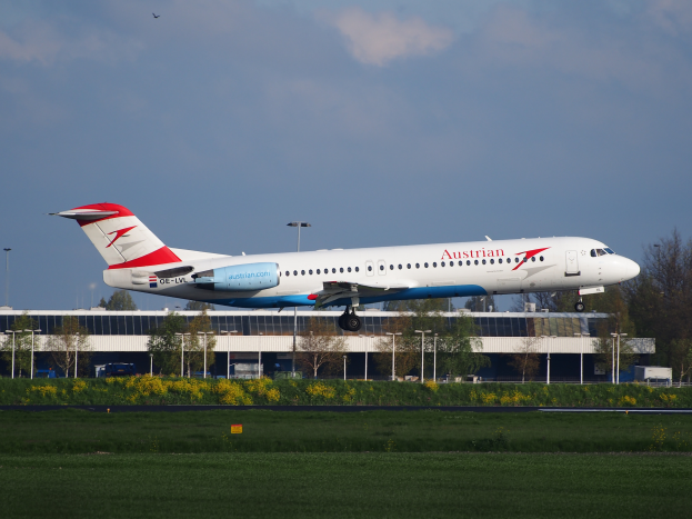 Ein Airbus A320-200 der Austrian Airlines startet am Frankfurter Flughafen, umgeben von grünem Gras, blühenden Pflanzen, Bäumen, Laternen und einem Gebäude unter einem bewölkten Himmel.