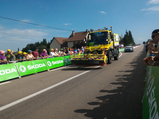Ein gelber Lkw fährt auf einer Straße neben einer Menge Menschen, einige tragen Mützen und Schutzbrillen, mit Schildern und Text an den Seiten der Straße, Bäumen, Gebäuden mit Fenstern und einem bewölkten Himmel im Hintergrund.