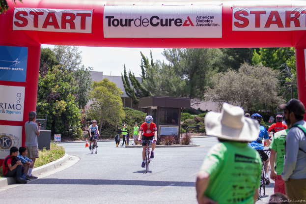 Eine belebte Straßenszene mit Menschen auf Fahrrädern, einige sitzen auf einem Gehweg und andere stehen auf der Straße, mit einer Hütte und umliegenden Pflanzen und Bäumen im Hintergrund.