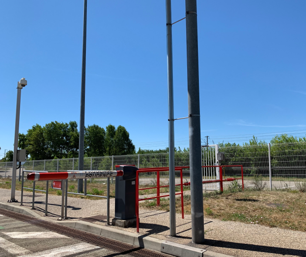 Ein Bahnhof mit einem roten und weißen Schild, umgeben von Pfählen, einem Metallzaun, Bäumen, Pflanzen, Gras, einem Straßenpfahl, Drähten und einem bewölkten Himmel, mit einer zentralen Schranke zum Schutz vor dem Betreten.