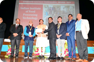 Eine Gruppe von Männern auf einer Bühne mit einem Zertifikat und einem Bildschirm im Hintergrund, der 'Indian Institute of Food Processing Technology Awards 2019' anzeigt, sowie Blumenschmuck.