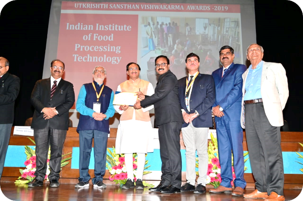 Eine Gruppe von Männern auf einer Bühne mit einem Zertifikat und einem Bildschirm im Hintergrund, der 'Indian Institute of Food Processing Technology Awards 2019' anzeigt, sowie Blumenschmuck.