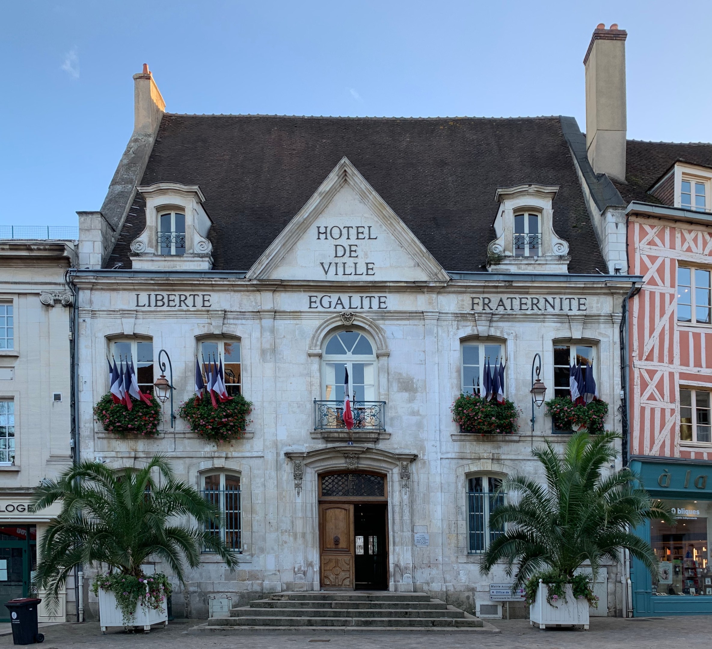 Das Rathaus in Saint-Emilio, Frankreich, ein Gebäude mit Fenstern, Türen und Text an der Wand, umgeben von Pflanzen mit Blumen, Bäumen, einem Müllcontainer und einer Treppe unter einem bewölkten Himmel.