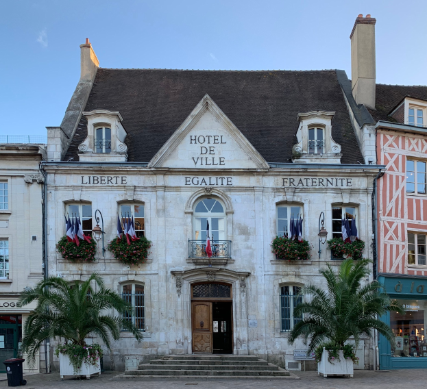 Das Rathaus in Saint-Emilio, Frankreich, ein Gebäude mit Fenstern, Türen und Text an der Wand, umgeben von Pflanzen mit Blumen, Bäumen, einem Müllcontainer und einer Treppe unter einem bewölkten Himmel.