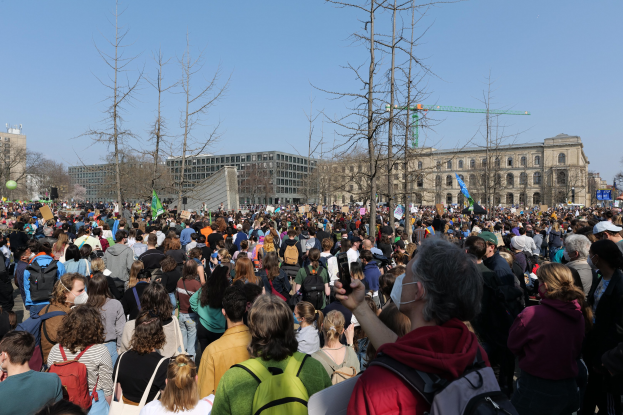 Große Menschenmenge bei einer Klimaprotestveranstaltung vor einem Gebäude mit Bäumen und einem klaren Himmel im Hintergrund, viele halten Schilder und tragen Taschen.