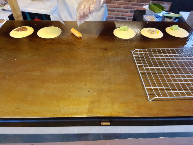 Eine Person steht in einer Küche und bereitet Essen auf einem Grill vor einem Tisch mit verschiedenen Lebensmitteln und einer Backsteinwand im Hintergrund.