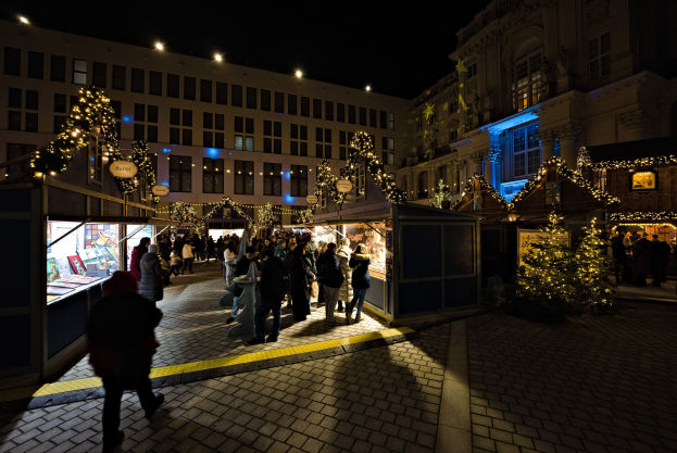 Ein lebendiger Weihnachtsmarkt in einer nächtlichen Stadt mit Menschen um hell erleuchtete Stände und geschmückte Gebäude im Hintergrund unter einem dunklen Himmel.