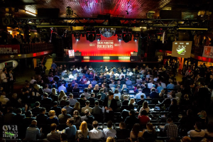 Eine große Menschenmenge sitzt auf Stühlen vor einer Bühne mit Bildschirmen und Plakaten, die von Scheinwerfern beleuchtet wird und von Geländern umgeben ist und gespannt auf den Beginn eines Online-Marketing-Rockstars-Events wartet, mit einem Wasserzeichen in der linken unteren Ecke.