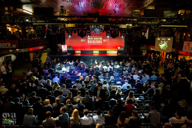 Eine große Menschenmenge sitzt auf Stühlen vor einer Bühne mit Bildschirmen und Plakaten, die von Scheinwerfern beleuchtet wird und von Geländern umgeben ist und gespannt auf den Beginn eines Online-Marketing-Rockstars-Events wartet, mit einem Wasserzeichen in der linken unteren Ecke.