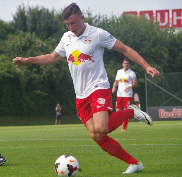 Ein Mann in einem Fußballtrikot schießt einen Ball auf einem grünen Feld mit Bäumen und einem klaren blauen Himmel im Hintergrund, mit einem Schild mit der Aufschrift "RB Leipzig" dahinter.