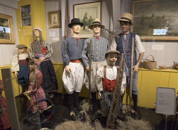 Eine Gruppe von Schaufensterpuppen in historischer Kleidung steht in einem Raum mit trockenem Gras auf dem Boden, einem Textschild, Fotorahmen an den Wänden, einem Schrank mit Gegenständen und einem Korb und anderen Gegenständen auf einer Plattform.