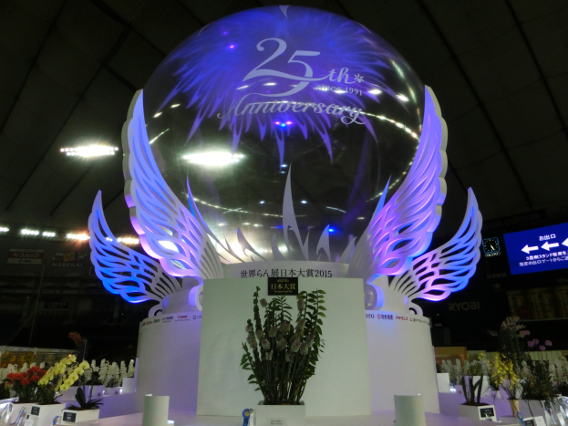 Ein großer Kristallball mit Flügeln oben drauf, umgeben von Blumensträußen, Namensschildern und anderen Gegenständen auf dem Boden, mit Tafeln, Lichtern und einem Bildschirm im Hintergrund bei einer festlichen 25-jährigen Jubiläumsfeier.