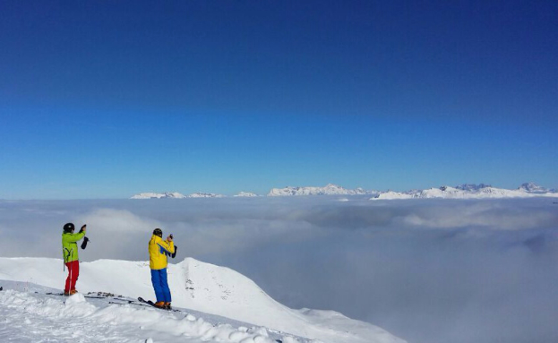 Zwei Personen auf Snowboards in einer verschneiten Landschaft, die Gegenstände halten, mit Bergen und Himmel im Hintergrund.