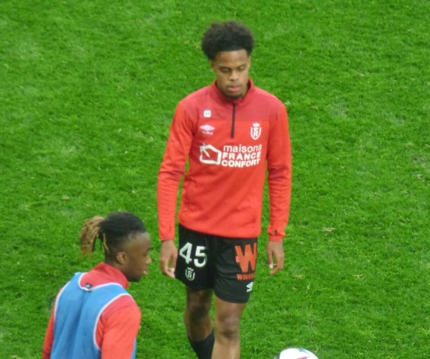 Zwei junge Männer stehen auf einem Fußballfeld nebeneinander mit einem Ball zu ihren Füßen, einer trägt eine Fußballuniform.