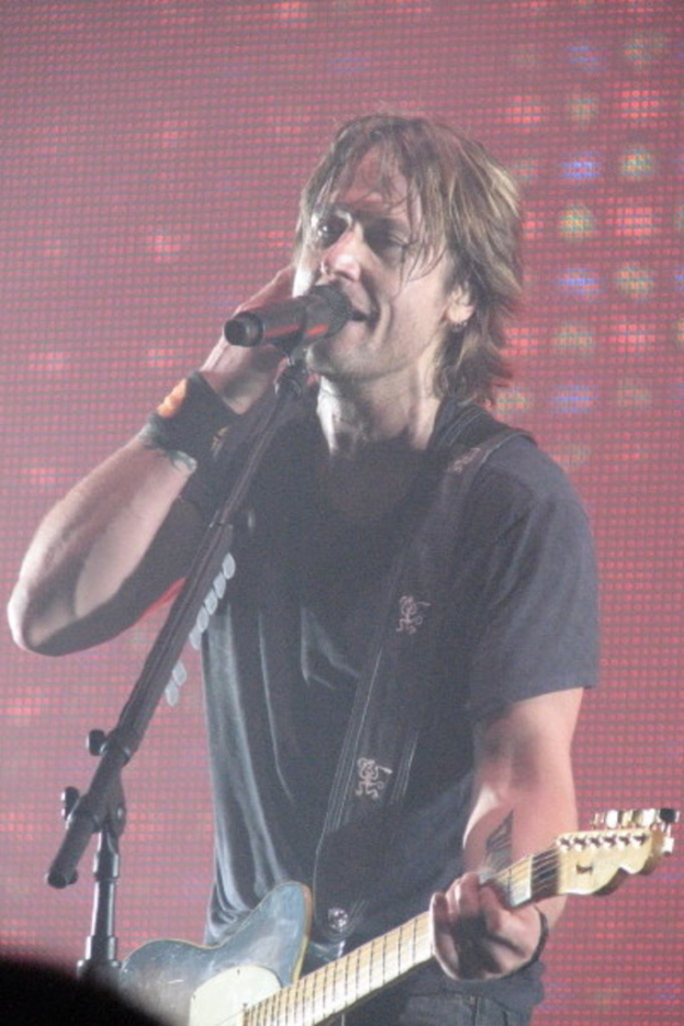 Ein Mann in einem T-Shirt singt in ein Mikrofon und hält eine Gitarre, während im Hintergrund ein Bildschirm oder eine Tafel zu sehen ist.