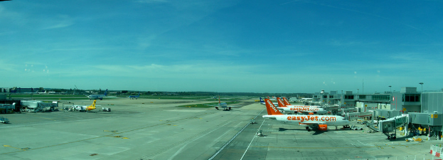 Eine Flughafen-Szene mit Flugzeugen, Gebäuden, Polen, Fahrzeugen und Bäumen unter einem Himmel mit einer Straße und einem Zaun im Vordergrund.