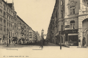 Schwarz-weiß-Fotografie einer Stadtstraße in Charlottenburg, Deutschland, mit Gebäuden, Straßenlaternen, Lampen, Namensschildern, Kraftfahrzeugen, Fußgängern und Gittern unter einem klaren Himmel.