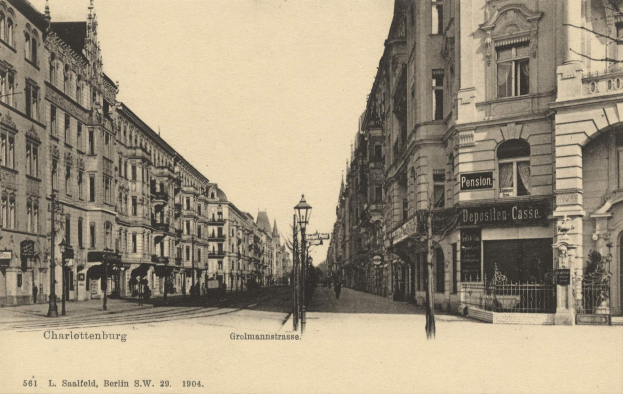 Schwarz-weiß-Fotografie einer Stadtstraße in Charlottenburg, Deutschland, mit Gebäuden, Straßenlaternen, Lampen, Namensschildern, Kraftfahrzeugen, Fußgängern und Gittern unter einem klaren Himmel.