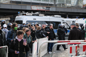 Eine Gruppe von Menschen steht vor Polizeifahrzeugen, einige tragen Helme und halten Telefone, mit Barrikaden im Vordergrund und einer Brücke mit Geländern und Gebäuden im Hintergrund in Berlin, Deutschland.