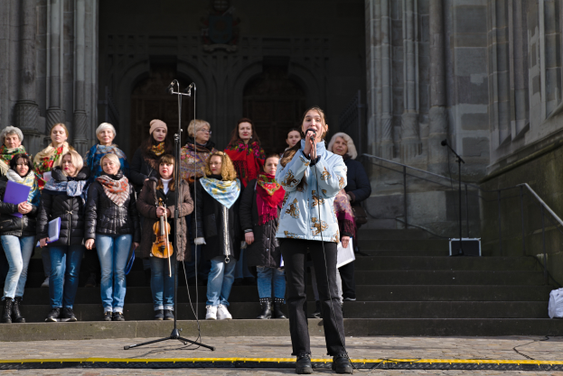 Eine Gruppe von Menschen steht vor einem Gebäude, wobei eine Frau in der Mitte ein Mikrofon hält und singt, umgeben von Mikrofonen mit Ständern, Musikinstrumenten, Treppen mit Geländern, einer Wand mit Säulen und einer Tür auf der rechten Seite.