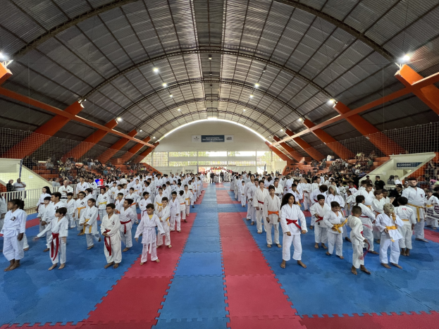 Eine große Gruppe von Menschen auf einer blauen und roten Matte in einer Turnhalle mit Zuschauern auf Treppen und Texttafeln im Hintergrund, die auf ein Taekwondo-Event hinweisen.