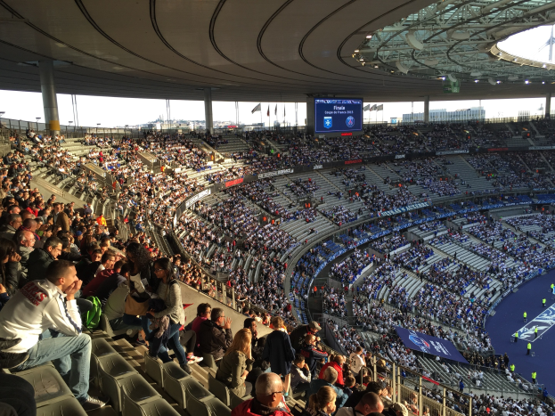 Große Menschenmenge in einem Stadion bei einem Fußballspiel, mit einer Bühne, Fahnen, Stangen und einem Bildschirm im Hintergrund.