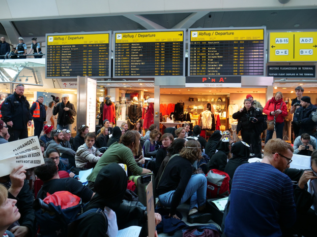 Eine große Gruppe von Menschen, die auf Stühlen und Stehplätzen in einem Flughafen während einer Protestaktion sitzen, mit einigen, die Taschen und Papiere halten, Hintergrundtafeln mit Text, Schaufensterpuppen in Kleidern und Deckenbeleuchtung.