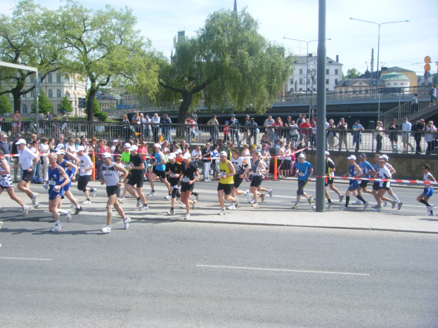 Gruppe von Menschen, die einen Marathon auf einer Straße mit Absperrungen, Zuschauern und städtischer Umgebung unter einem bewölkten Himmel laufen.