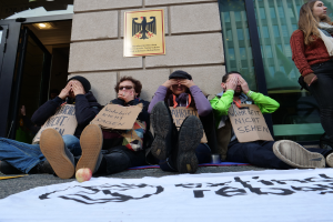 Eine Gruppe von Menschen sitzt vor einem Gebäude auf dem Boden und hält Schilder in den Händen, mit einem Banner und einem Apfel auf der Straße, und eine Frau steht rechts daneben und sieht besorgt aus.
