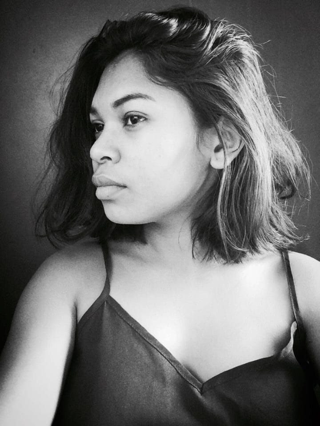 Eine selbstbewusste Frau in einem schwarzen Kleid posiert in einem Schwarz-Weiß-Foto, ihr glatter Bob und dezent Make-up unterstreichen ihre selbstbewusste Haltung vor einer Wand.