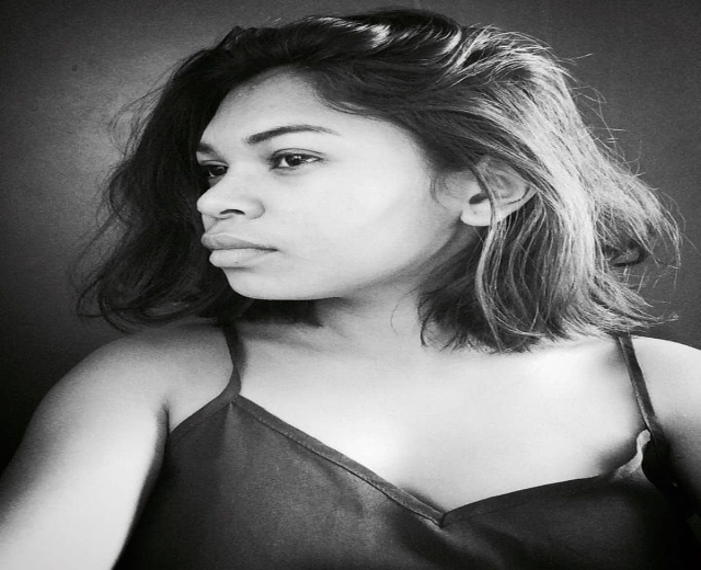 Eine selbstbewusste Frau in einem schwarzen Kleid posiert in einem Schwarz-Weiß-Foto, ihr glatter Bob und dezent Make-up unterstreichen ihre selbstbewusste Haltung vor einer Wand.