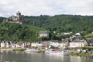 Eine malerische Aussicht auf den Rhein in Deutschland, mit einer Burg auf einem Hügel im Hintergrund, Booten auf dem Fluss und Fahrzeugen auf einer nahen Straße sowie einem bewölkten Himmel.
