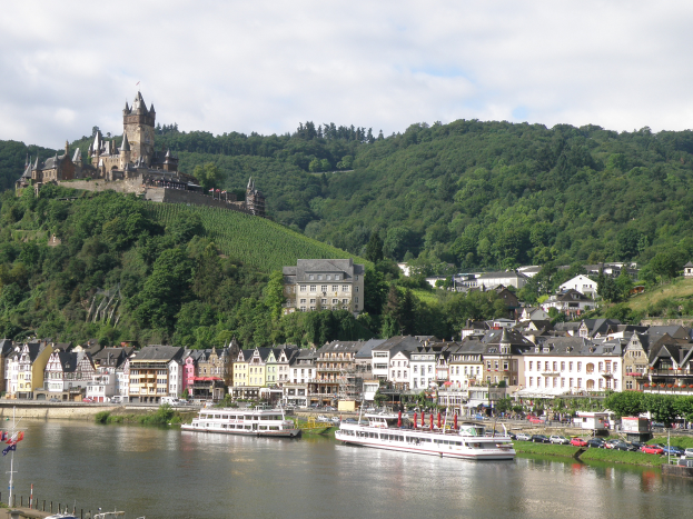 Eine malerische Aussicht auf den Rhein in Deutschland, mit einer Burg auf einem Hügel im Hintergrund, Booten auf dem Fluss und Fahrzeugen auf einer nahen Straße sowie einem bewölkten Himmel.