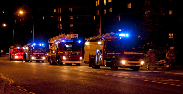 Feuerwehrlöschfahrzeuge sind nachts auf der Straße unterwegs, mit Gebäuden und Laternen im Hintergrund und zwei Personen auf der rechten Seite.