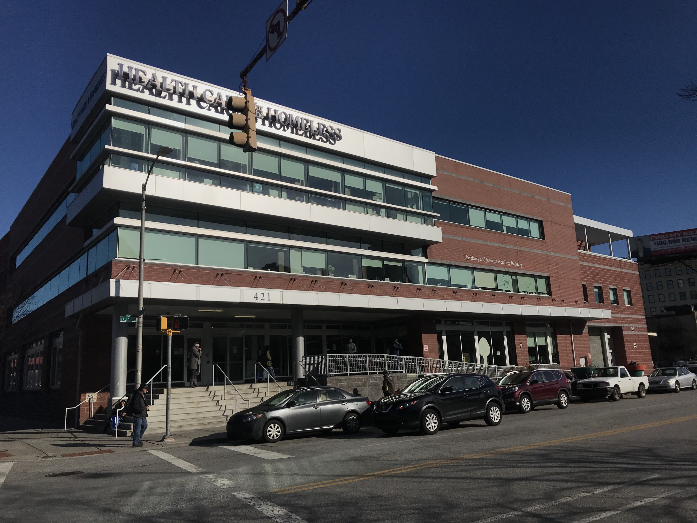 Großes Gebäude mit der Aufschrift "New York City Health Care Homeless Center" mit parkenden Autos, Fußgängern, Straßenmöbeln und Bäumen unter einem klaren blauen Himmel.