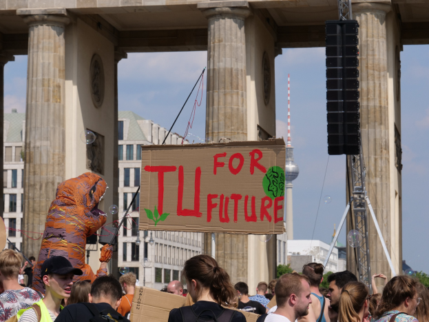 Eine Gruppe von Menschen steht vor dem Brandenburger Tor in Berlin, Deutschland, wobei einige Taschen tragen und einer im Kostüm mit einer Tafel in der Hand zu sehen ist, vor einem Hintergrund von Gebäuden, Bäumen, einem Turm und einem bewölkten Himmel.