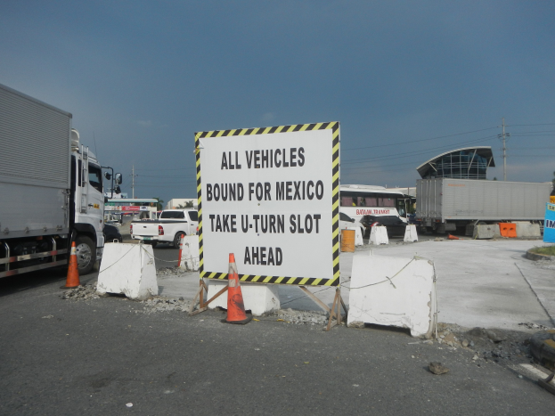Ein Lkw parkt auf einem Parkplatz neben einem Schild mit der Aufschrift "Alle Fahrzeuge Richtung Mexiko", mit verstreuten Konen, Brettern und anderen Gegenständen und im Hintergrund Gebäude, Bäume, Pfosten und einen klaren blauen Himmel.