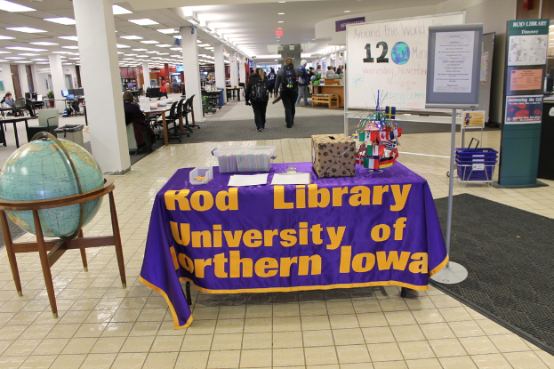 Eine Ausstellung mit einem Tisch, auf dem verschiedene Landesflaggen, eine Schachtel, Papiere und ein Banner liegen, eine Kugel links daneben, ein Anschlagbrett rechts daneben mit ein paar Leuten in der Nähe und mehreren Tischen und Stühlen ganz links.