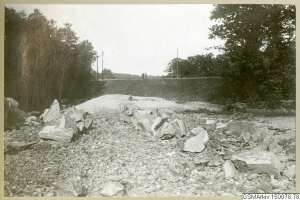 Schwarz-weiß-Foto einer Schotterstraße mit verstreuten Steinen, Bäumen, Strommasten, einer Wand im Hintergrund und dem Himmel darüber, mit einer Liste von Orten und Routen unten.