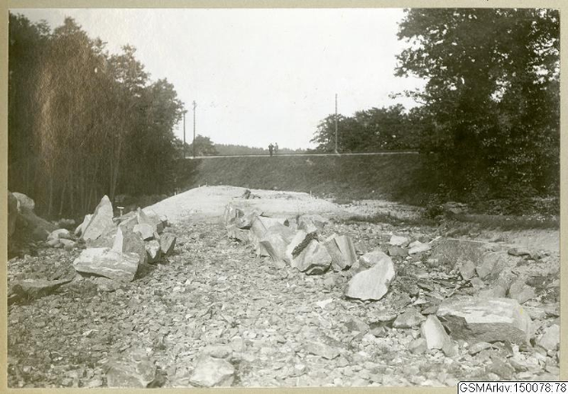 Schwarz-weiß-Foto einer Schotterstraße mit verstreuten Steinen, Bäumen, Strommasten, einer Wand im Hintergrund und dem Himmel darüber, mit einer Liste von Orten und Routen unten.