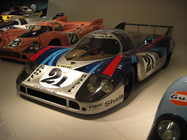 Ein Porsche 917K Rennwagen auf einer Museumsausstellung, umgeben von anderen Sportwagen und Menschen, mit einer Wand und informativem Text im Hintergrund.